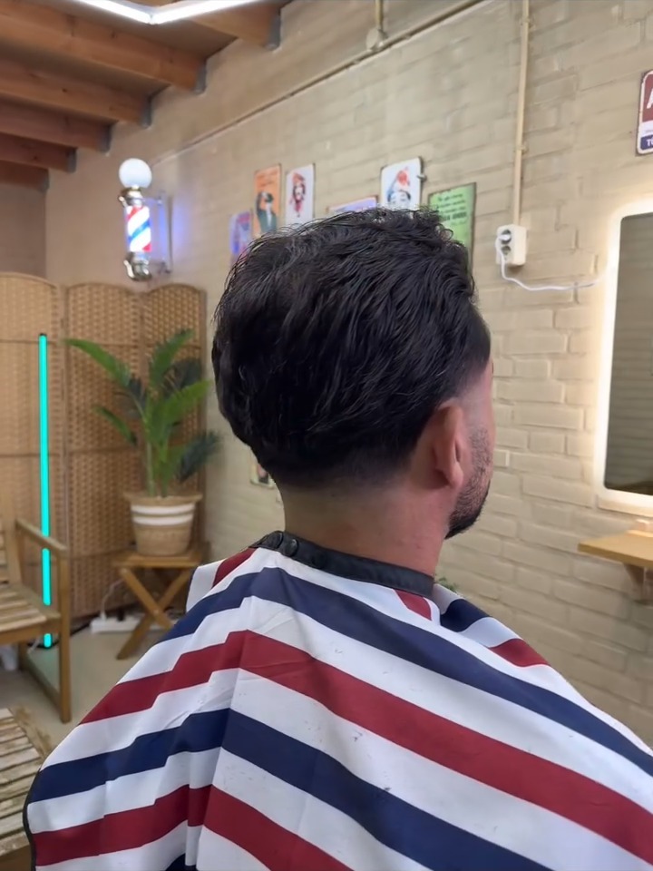 Klant in een rood-wit-blauwe kapmantel van achteren, met de barberpaal en een plant in de studio