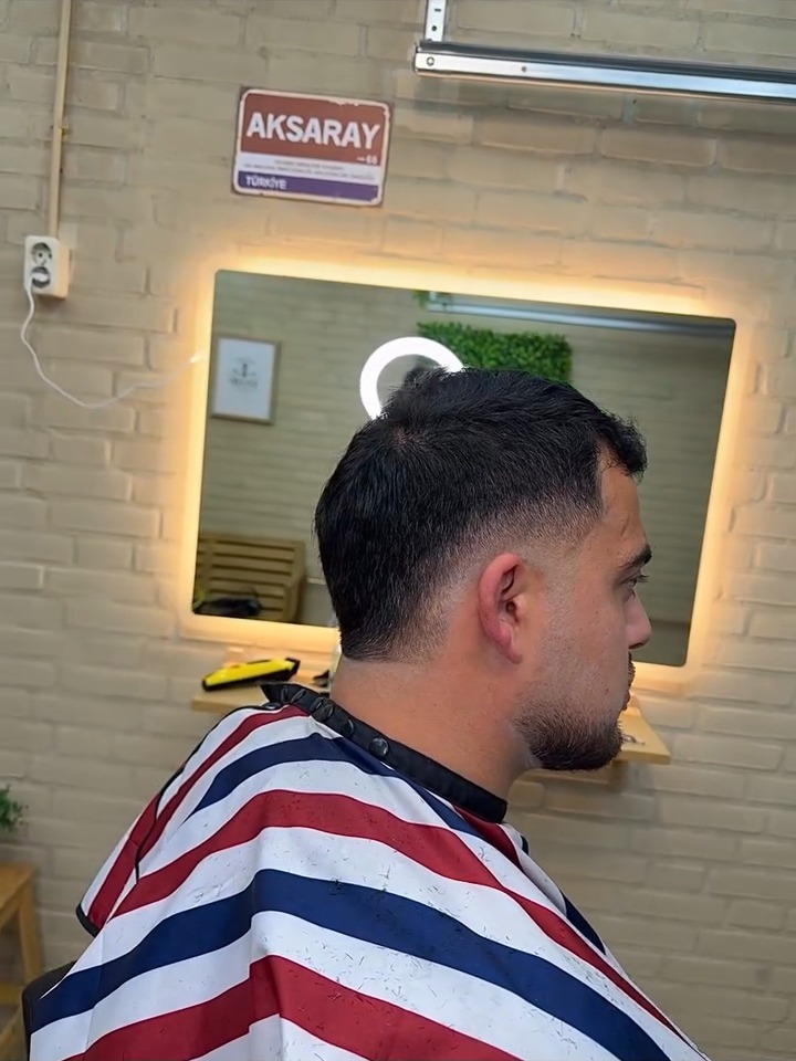 Klant met een korte fade in een gestreepte kapmantel, voor de verlichte spiegel in de studio